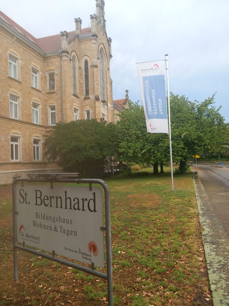 Gepflegte Grünfläche vor dem Bildungshaus St. Bernhard mit Hinweisschild und Fahne